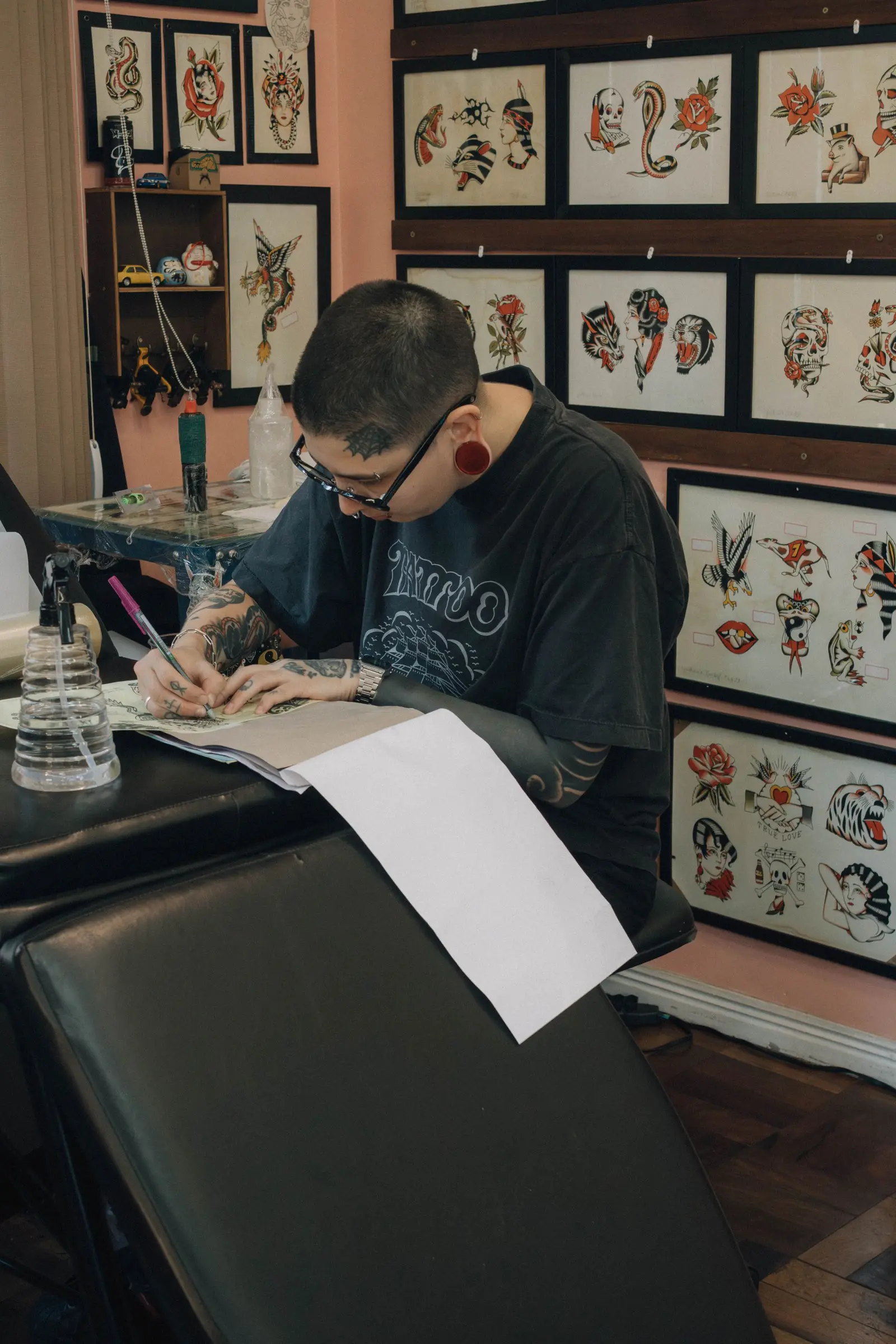 Tattoo artist drawing at a desk in front of a wall of traditional flash